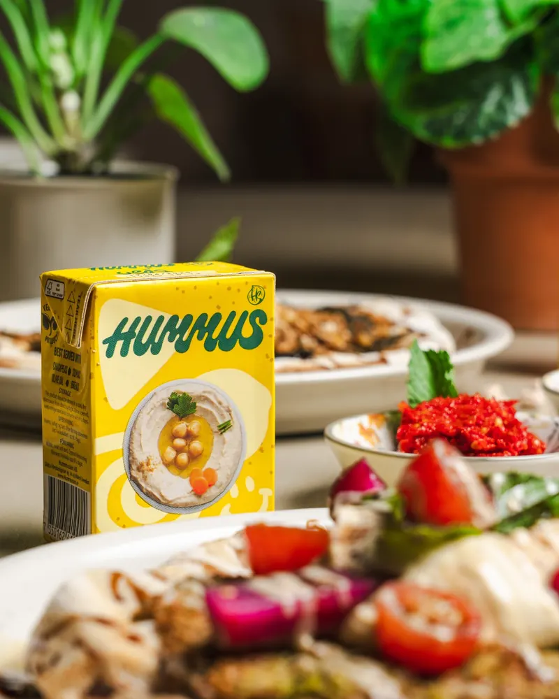 A carton of Hummusson's packaged hummus on the table, with plates of food in the background